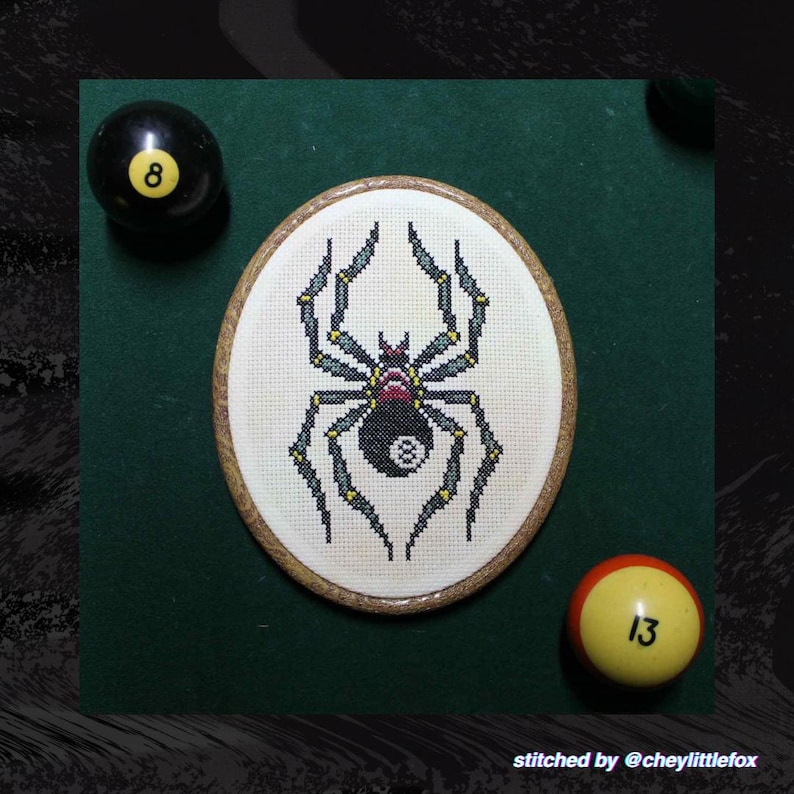 Puede incluir: Un bordado de punto de cruz ovalado con una ara&ntilde;a con un dise&ntilde;o de bola 8. La ara&ntilde;a es negra, gris y amarilla sobre un fondo blanco. Tambi&eacute;n hay dos bolas de billar, una negra con un 8 y otra amarilla con un 13.