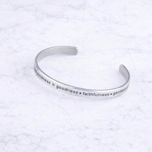 May include: A silver-colored cuff bracelet with the words "kindness, goodness, faithfulness, gentleness" engraved on the outside. The bracelet is open-ended and sits on a white surface.