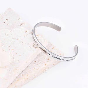 Può includere: Un bracciale a polsino color argento con le parole "kindness, goodness, faithfulness, gentleness" incise. Il bracciale aperto è appoggiato su una superficie strutturata bianco sporco. Design semplice ed elegante.