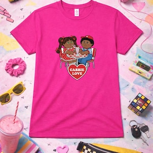 Op de afbeelding: Felroze t-shirt met een cartoonillustratie van twee poppen die een milkshake delen aan een tafel. Het ontwerp bevat een rood hart met de tekst "CABBIE LOVE".