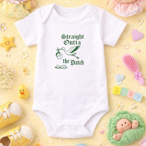 Puede incluir: Body de bebé blanco con el texto "Straight Outta the Patch" en verde. El diseño presenta una cigüeña que lleva un bebé. El body está rodeado de juguetes y accesorios para bebés, incluyendo muñecas, zapatos y bloques.