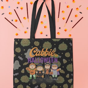 May include: Black tote bag with a Halloween-themed design. The bag features a pattern of pumpkins and ghosts, with the text "Cabbie Halloween" and a graphic of four costumed characters. The bag has black handles and is set against a pink background with candy corn and decorative elements.