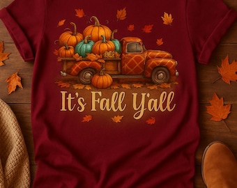 Puede incluir: Camiseta burdeos con un diseño otoñal. El diseño presenta una camioneta naranja vintage llena de calabazas naranjas y verde azulado, rodeada de hojas de otoño. El texto "It's Fall Y'all" está impreso en oro.