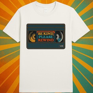 May include: White t-shirt with a retro VHS tape design. The tape is teal, orange, and yellow, with the words "BE KIND, PLEASE REWIND". A nostalgic design for film enthusiasts.