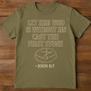 May include: Olive green t-shirt with the text "LET HIM WHO IS WITHOUT SIN CAST THE FIRST STONE" and a stone graphic. The text "-JOHN 8:7" is below the graphic. The shirt has a crew neck and short sleeves.