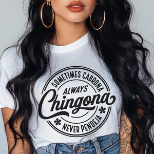 May include: A white t-shirt with a black circle graphic that says "Sometimes Cabrona Always Chingona Never Pendeja". The model is wearing a red and green bandana and large hoop earrings.