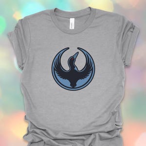May include: A heather gray t-shirt featuring a circular design with a stylized bird in shades of blue. The bird is depicted with outstretched wings within a blue circle. The shirt is made of a soft material.