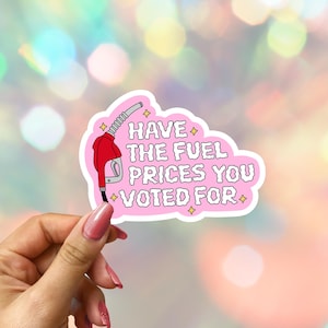 May include: A pink sticker with the text "Have the fuel prices you voted for" and a red gas pump graphic. The sticker has a white border and is held by a hand with pink nail polish.