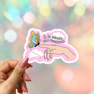 May include: A pink sticker featuring a butterfly with blue and yellow wings, perched on a finger. The sticker has the text "A la mierda sus fronteras" in black. The background is a bokeh of colorful lights.