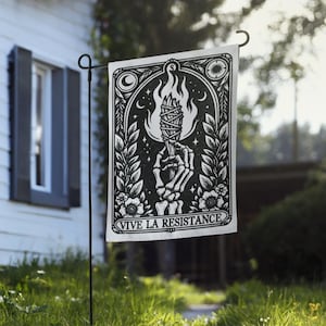 May include: A black and white garden flag with the words "VIVE LA RESISTANCE" at the bottom. The flag features a skeleton hand holding a torch with flames, surrounded by floral and celestial designs. The flag is displayed on a black metal stand in a grassy yard.