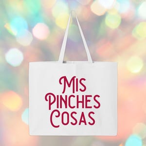 May include: White tote bag with red text that reads "MIS PINCHES COSAS". The bag has a rectangular shape with a handle. The background is a blurred mix of colorful bokeh lights.