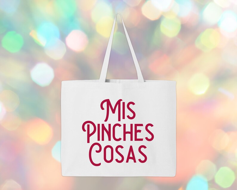 May include: White tote bag with red text that reads "MIS PINCHES COSAS". The bag has a rectangular shape with a handle. The background is a blurred mix of colorful bokeh lights.