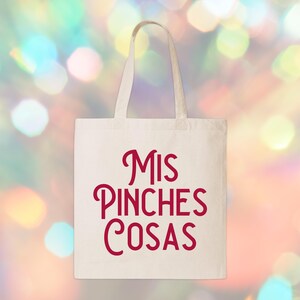 May include: A white canvas tote bag with the words "Mis Pinches Cosas" in red script. The bag has two handles and is set against a blurred background of colorful bokeh lights. The tote bag is a reusable shopping bag.