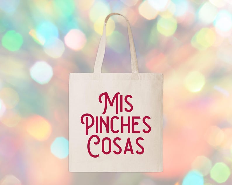 May include: A white canvas tote bag with the words "Mis Pinches Cosas" in red script. The bag has two handles and is set against a blurred background of colorful bokeh lights. The tote bag is a reusable shopping bag.