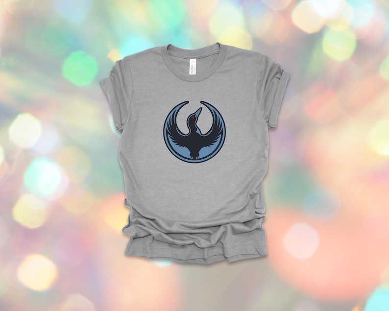 May include: A heather gray t-shirt featuring a circular design with a stylized bird in shades of blue. The bird is depicted with outstretched wings within a blue circle. The shirt is made of a soft material.