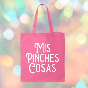 May include: A pink tote bag with the words "Mis Pinches Cosas" in white script. The bag has a rectangular shape with a handle. The background is a blurred mix of colorful bokeh lights.