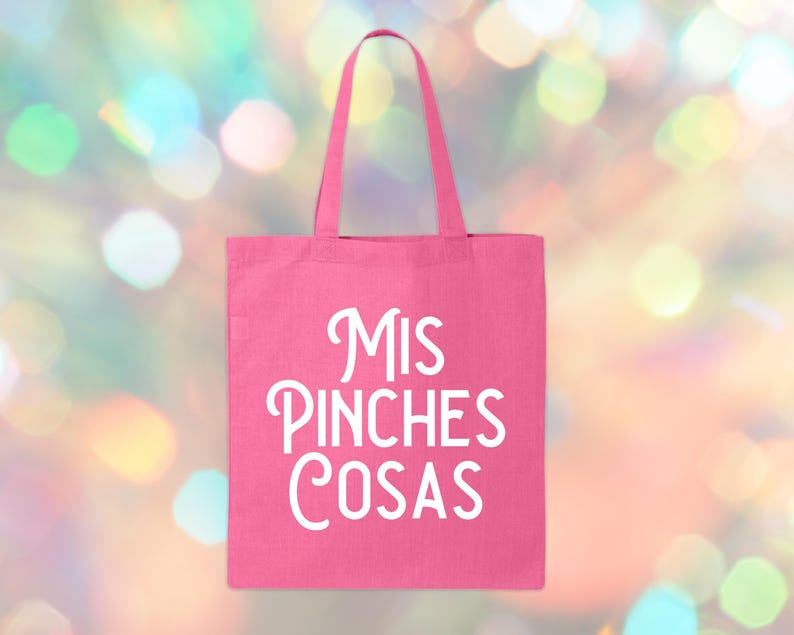 May include: A pink tote bag with the words "Mis Pinches Cosas" in white script. The bag has a rectangular shape with a handle. The background is a blurred mix of colorful bokeh lights.