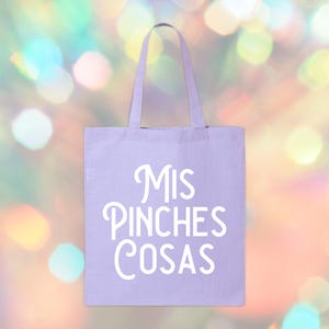 May include: A light purple tote bag with the words "Mis Pinches Cosas" in white script. The bag has two handles and is made of a canvas-like material. The background is a blurred mix of colorful bokeh lights.