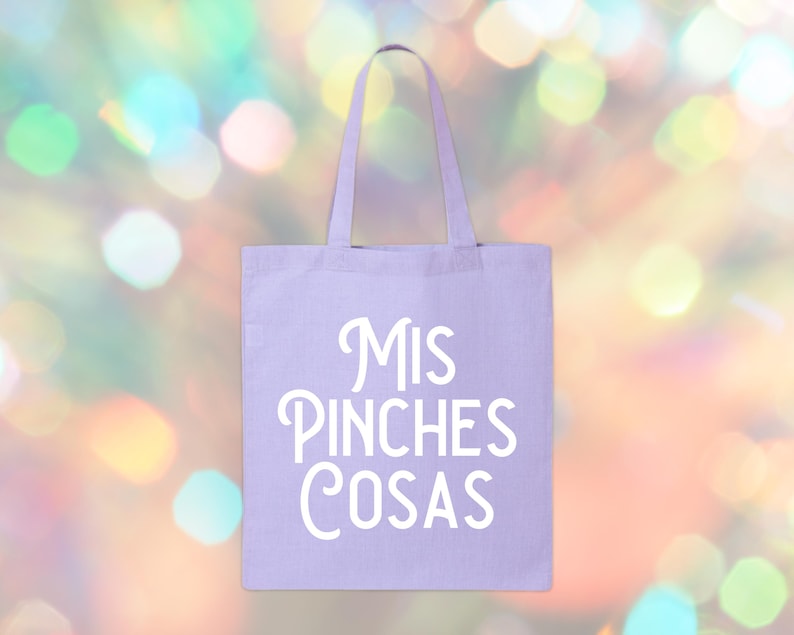 May include: A light purple tote bag with the words "Mis Pinches Cosas" in white script. The bag has two handles and is made of a canvas-like material. The background is a blurred mix of colorful bokeh lights.