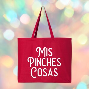 May include: A red tote bag with the words "Mis Pinches Cosas" in white script. The bag has long handles and is made of a sturdy fabric. The background is a blurred mix of colorful lights.