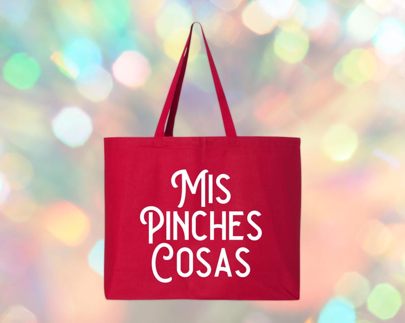 May include: A red tote bag with the words "Mis Pinches Cosas" in white script. The bag has long handles and is made of a sturdy fabric. The background is a blurred mix of colorful lights.