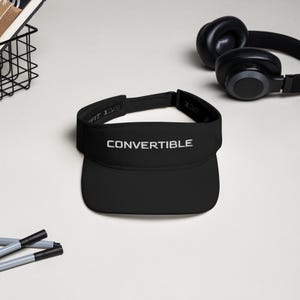 May include: Black visor with the word "CONVERTIBLE" embroidered in white. The visor has a curved brim and a strap to adjust the fit. A pair of black headphones are in the background.
