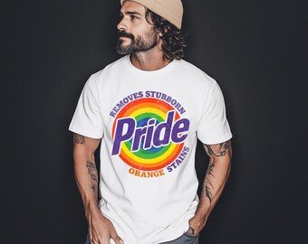 以下が含まれることがあります： 虹色のPrideグラフィックがプリントされた白いTシャツ。グラフィックには、紫色の「Pride」の文字、虹、そして「Removes Stubborn Orange Stains」のテキストが含まれています。Tシャツはクルーネックです。