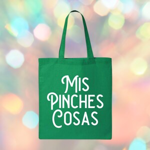 May include: A green tote bag with white text that reads "Mis Pinches Cosas". The bag has a rectangular shape with a handle at the top. The background is a blurred mix of colorful lights.