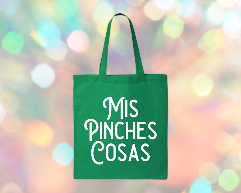 May include: A green tote bag with white text that reads "Mis Pinches Cosas". The bag has a rectangular shape with a handle at the top. The background is a blurred mix of colorful lights.