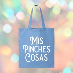 May include: A light blue tote bag with white text that reads "Mis Pinches Cosas". The bag has two handles and is made of a sturdy fabric. The background is a colorful bokeh effect.
