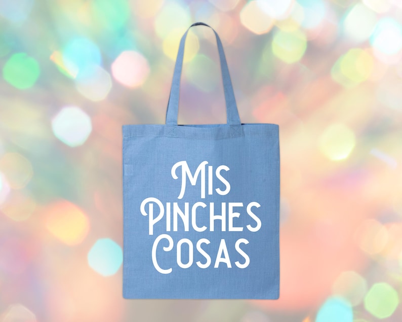 May include: A light blue tote bag with white text that reads "Mis Pinches Cosas". The bag has two handles and is made of a sturdy fabric. The background is a colorful bokeh effect.