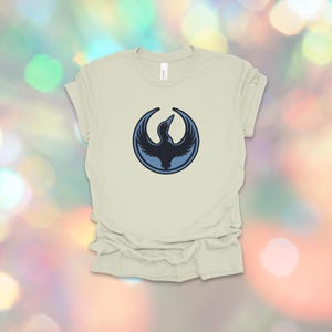 May include: A light beige t-shirt featuring a circular emblem with a dark blue bird design. The bird is depicted with outstretched wings, set against a lighter blue circle. The shirt is made of soft material.
