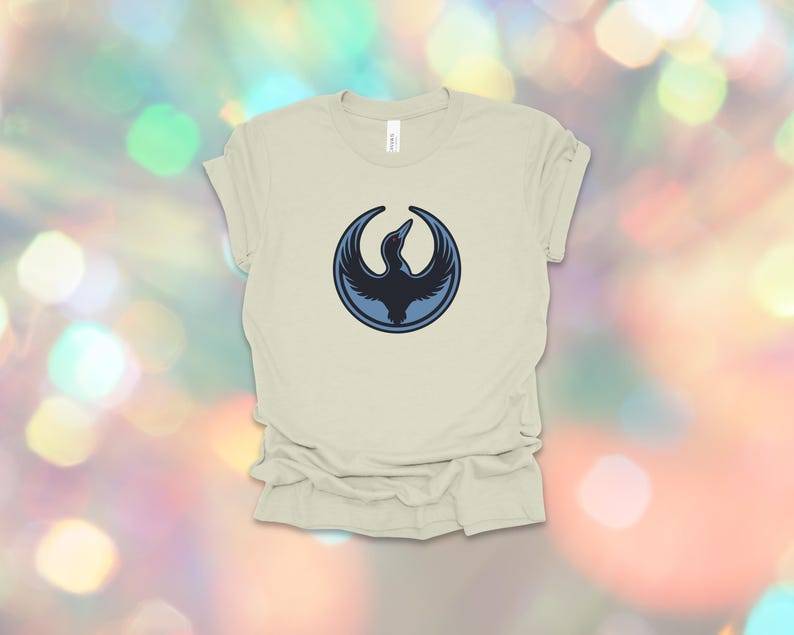 May include: A light beige t-shirt featuring a circular emblem with a dark blue bird design. The bird is depicted with outstretched wings, set against a lighter blue circle. The shirt is made of soft material.