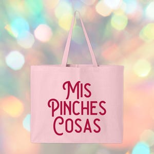 May include: A light pink tote bag with the phrase "MIS PINCHES COSAS" in burgundy script. The bag has a rectangular shape with two long handles. The background is a blurred mix of colorful bokeh lights.