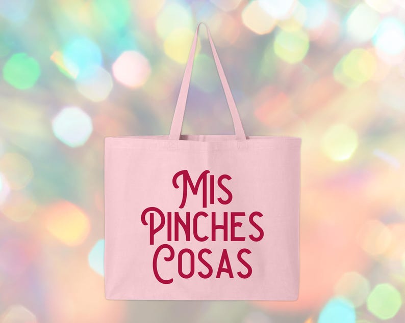 May include: A light pink tote bag with the phrase "MIS PINCHES COSAS" in burgundy script. The bag has a rectangular shape with two long handles. The background is a blurred mix of colorful bokeh lights.
