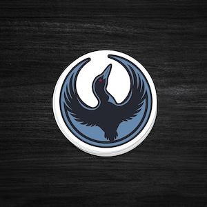 May include: A round sticker featuring a stylized bird with outstretched wings. The bird is primarily dark blue with light blue accents and a red eye, set against a white background within a blue border. The sticker is on a dark wood surface.