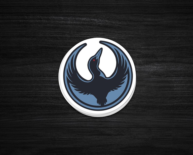 May include: A round sticker featuring a stylized bird with outstretched wings. The bird is primarily dark blue with light blue accents and a red eye, set against a white background within a blue border. The sticker is on a dark wood surface.