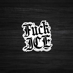 May include: A white sticker with the words "Fuck ICE" in a gothic font. The sticker is on a dark wood grain surface. The text is in bold, black letters.