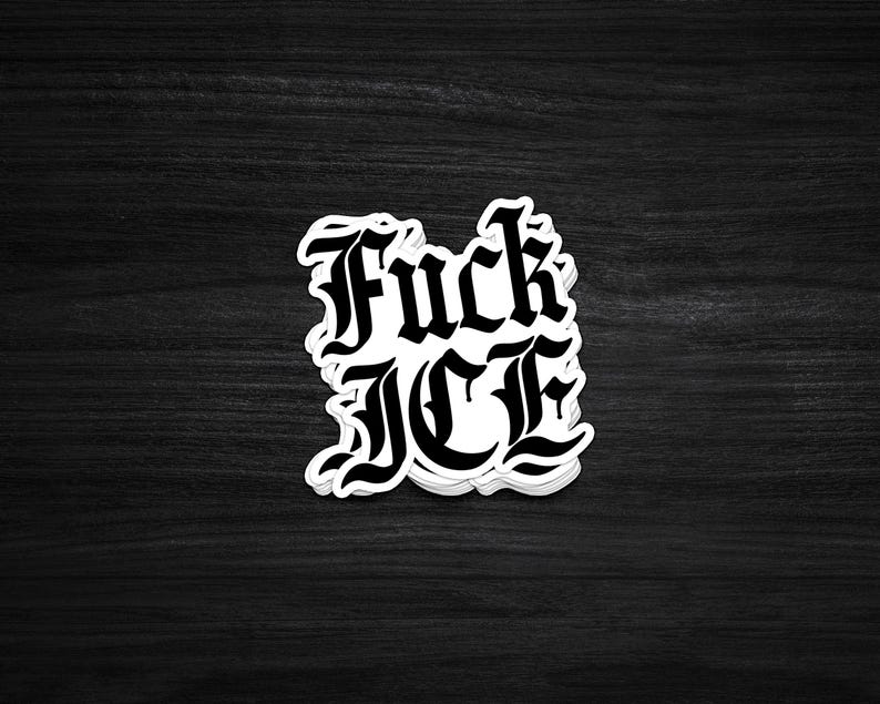 May include: A white sticker with the words "Fuck ICE" in a gothic font. The sticker is on a dark wood grain surface. The text is in bold, black letters.