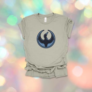 May include: A light olive green t-shirt featuring a circular design with a dark blue bird graphic. The bird is in flight, with its wings spread. The shirt is made of a soft material and has a classic crew neck.