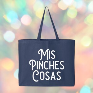 May include: Navy blue tote bag with white text that reads "Mis Pinches Cosas". The bag has a rectangular shape with a handle at the top. The background is a colorful bokeh effect.