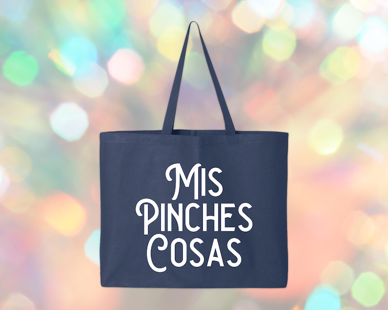 May include: Navy blue tote bag with white text that reads "Mis Pinches Cosas". The bag has a rectangular shape with a handle at the top. The background is a colorful bokeh effect.