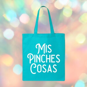May include: A turquoise tote bag with white text that reads "Mis Pinches Cosas." The bag has a rectangular shape with a handle at the top. The background is a blurred bokeh of colorful lights.