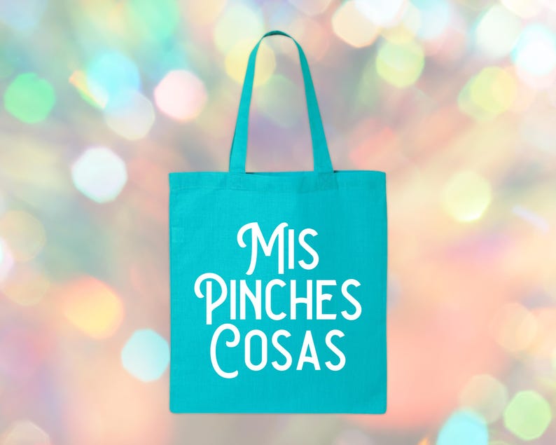 May include: A turquoise tote bag with white text that reads "Mis Pinches Cosas." The bag has a rectangular shape with a handle at the top. The background is a blurred bokeh of colorful lights.
