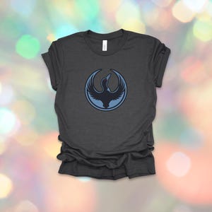 May include: Dark gray t-shirt featuring a circular emblem with a stylized bird design in shades of blue. The bird is depicted with outstretched wings, set against a light blue outline. The shirt is made of soft material.
