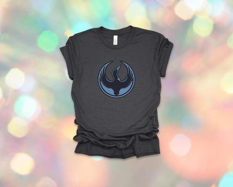 May include: Dark gray t-shirt featuring a circular emblem with a stylized bird design in shades of blue. The bird is depicted with outstretched wings, set against a light blue outline. The shirt is made of soft material.