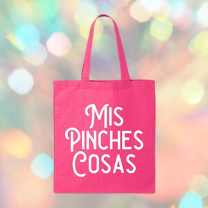 May include: A bright pink tote bag with white text that reads "Mis Pinches Cosas". The bag has a rectangular shape with a handle. The background is a colorful bokeh effect.