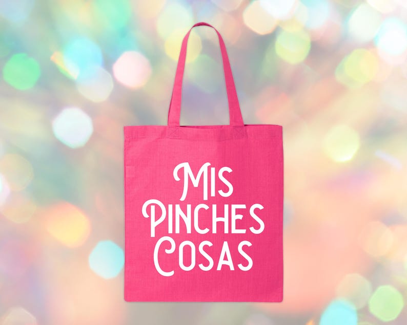May include: A bright pink tote bag with white text that reads "Mis Pinches Cosas". The bag has a rectangular shape with a handle. The background is a colorful bokeh effect.