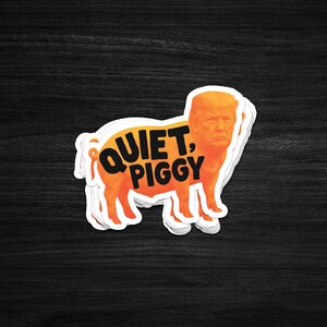 Pode incluir: Um autocolante com um porco laranja com o rosto de um homem. O texto "QUIET, PIGGY" est&aacute; impresso em laranja no corpo do porco. O autocolante tem uma borda branca e est&aacute; colocado sobre uma superf&iacute;cie de madeira escura.