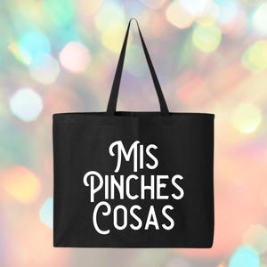 May include: A black tote bag with the words "MIS PINCHES COSAS" in white script. The bag has a rectangular shape with a handle extending from the top. The background is a colorful bokeh effect.